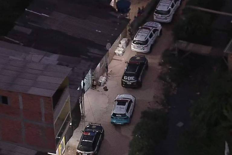 A imagem mostra uma operação policial em uma área urbana， com várias viaturas estacionadas em uma rua estreita. As viaturas são predominantemente brancas com detalhes em azul e preto， e algumas têm a palavra 039;Polícia039; visível. O ambiente é composto por casas de construção simples， com telhados de metal e paredes de tijolos expostos. Há também algumas pessoas visíveis na cena.