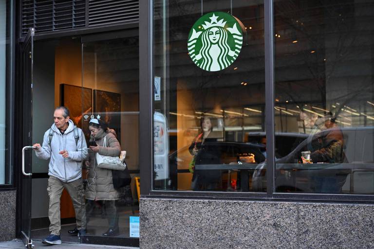 A imagem mostra a entrada de uma cafeteria Starbucks. Um homem está saindo pela porta， enquanto uma mulher está dentro， próxima à janela. O logotipo verde da Starbucks é visível na parte superior da imagem. O ambiente parece moderno e acolhedor， com mesas e cadeiras visíveis através da janela.