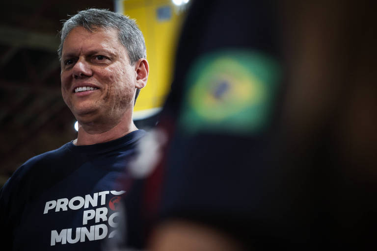 A imagem mostra Tarcísio de Freitas， homem grisalho， sorrindo， vestindo uma camiseta preta com a frase 039;PRONTO PRO MUNDO039;. Ao fundo， é possível ver uma bandeira do Brasil em um uniforme. O ambiente parece ser um evento ou reunião.