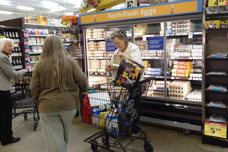 A imagem mostra um supermercado com prateleiras de produtos refrigerados. Um homem está pegando um item de uma prateleira， enquanto duas mulheres estão próximas a ele， uma delas com cabelo longo e grisalho. Um carrinho de compras está ao lado do homem， cheio de produtos.