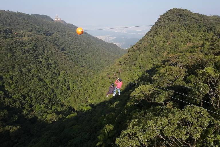 A imagem mostra duas pessoas em uma tirolesa， suspensas sobre uma densa vegetação verde em uma montanha. Ao fundo， é possível ver uma cidade e uma estrutura arquitetônica no topo da montanha. O céu está claro e a iluminação é natural， destacando a beleza da paisagem.