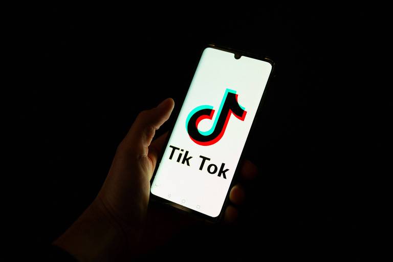 A imagem mostra um smartphone sendo segurado por uma mão， exibindo o logotipo do TikTok em uma tela clara. O logotipo é composto por um símbolo de nota musical em cores vermelha e azul， com o texto 039;Tik Tok039; abaixo dele. O fundo da imagem é escuro.