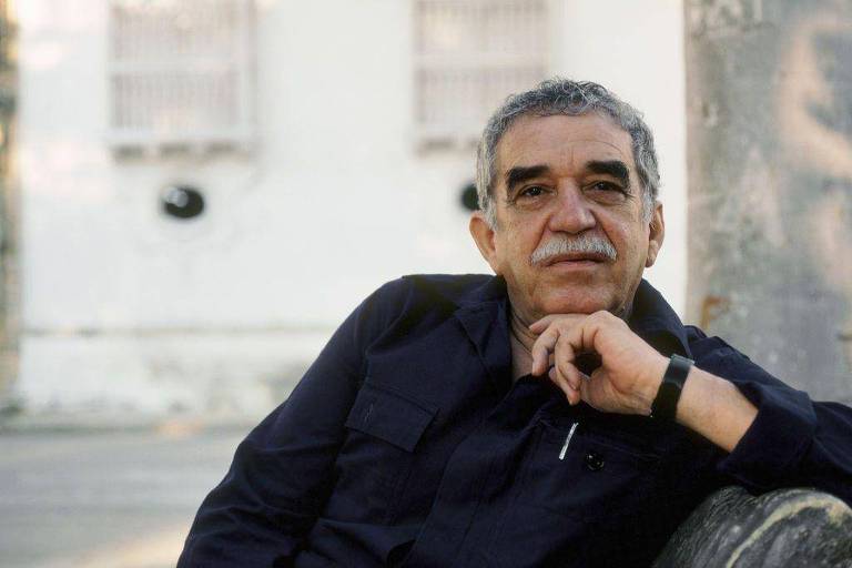 A imagem mostra Gabriel Garcia Marquez. Um homem de pele clara e cabelo grisalho está sentado， apoiando o queixo na mão. Ele usa uma camisa escura e tem um olhar contemplativo. Ao fundo， há uma parede clara com janelas e marcas de desgaste visíveis.