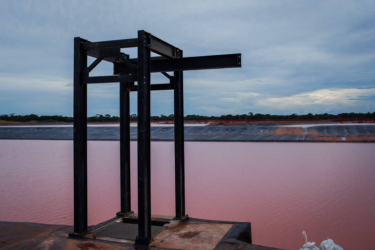 A imagem mostra uma estrutura metálica preta em primeiro plano, localizada em uma área próxima a uma lagoa de cor rosa. O céu está nublado, com nuvens cinzentas, e ao fundo é possível ver a extensão da lagoa rosa, que reflete a luz do céu. A estrutura parece ser um suporte ou equipamento, e há uma superfície de metal ao redor.