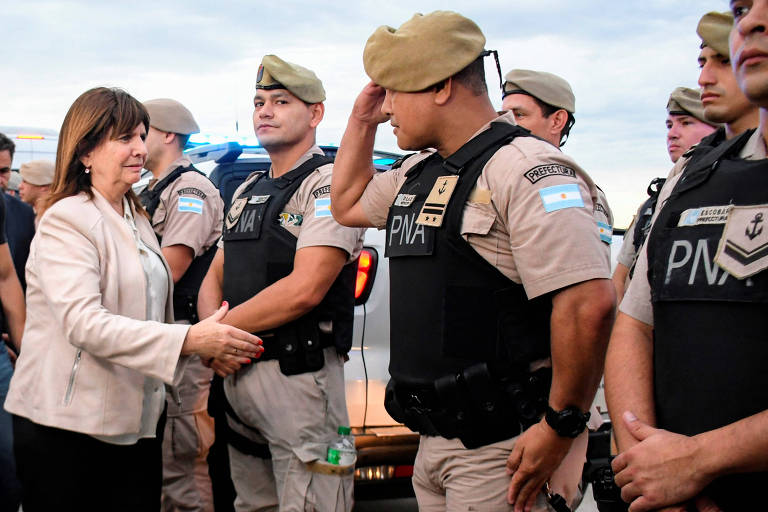 A imagem mostra uma mulher cumprimentando um policial em uniforme da Polícia Nacional Argentina. O policial está em posição de saudação， enquanto outros policiais em uniforme estão alinhados ao fundo. A mulher usa uma blusa clara e está estendendo a mão para o policial. O ambiente parece ser ao ar livre， com veículos da polícia visíveis ao fundo.