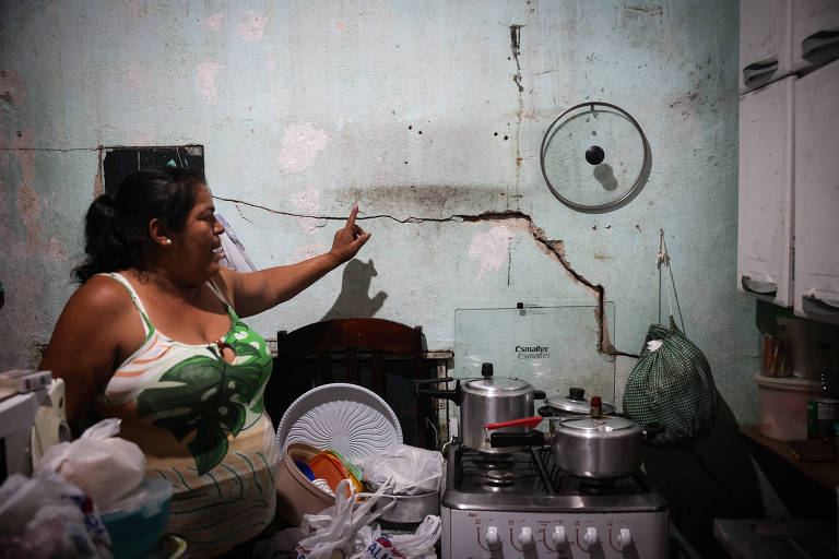 A imagem mostra uma mulher em uma cozinha com paredes desgastadas e uma fissura visível. Ela está apontando para a parede， que apresenta manchas e uma linha de rachadura. A cozinha é simples， com um fogão à gás e utensílios de cozinha visíveis. Há sacolas plásticas e outros objetos espalhados pela bancada.