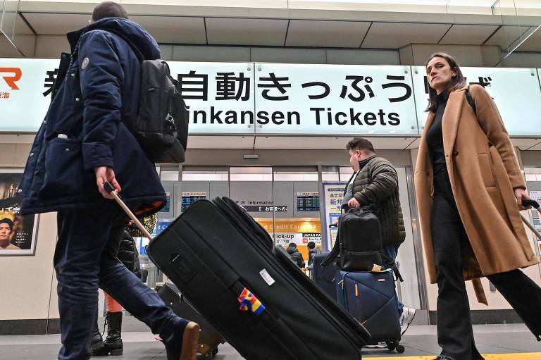 A imagem mostra uma estação de trem com pessoas caminhando. Um homem está puxando uma mala grande， enquanto uma mulher passa ao lado dele. Ao fundo， há um sinal que diz 039;Shinkansen Tickets039; em inglês e japonês. O ambiente parece movimentado， típico de uma estação de transporte.