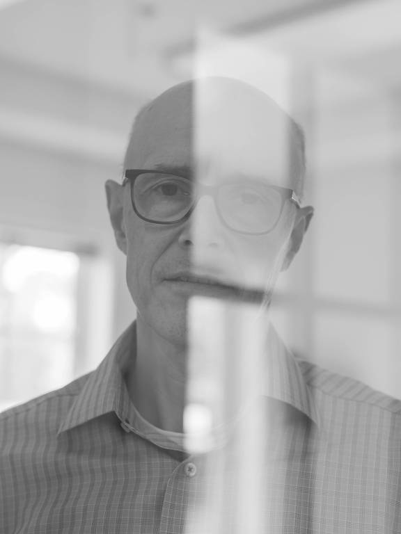 A imagem mostra um homem com cabelo calvo e óculos, olhando diretamente para a câmera. Ele está em um ambiente interno, com luz natural visível ao fundo. A foto é em preto e branco, e há um efeito de desfoque ou reflexo que obscurece parcialmente sua face.
