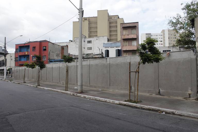 A imagem mostra uma rua urbana com um muro cinza ao fundo. À esquerda, há um prédio de cor vermelha e azul, enquanto à direita, edificações de cores neutras e um prédio mais alto em tons claros. Algumas árvores pequenas estão plantadas ao longo do muro