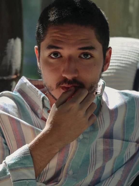 Um jovem está sentado em um sofá, olhando diretamente para a câmera. Ele tem cabelo curto e uma barba bem aparada. Está vestindo uma camisa de botão com listras em cores suaves, como azul, rosa e branco. O jovem está mordendo o dedo indicador, com uma expressão pensativa. Ao fundo, há uma luz suave que ilumina o ambiente.