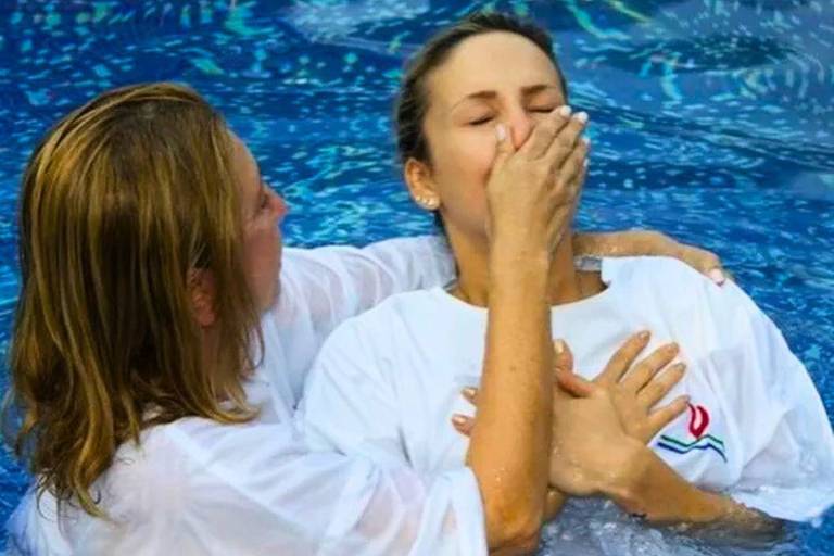 Duas mulheres estão em uma piscina. Uma mulher, com cabelo loiro e molhado, está segurando o rosto da outra mulher, que tem cabelo preso e está com os olhos fechados. Ambas estão vestidas com camisetas brancas. A água da piscina é azul e reflete a luz.