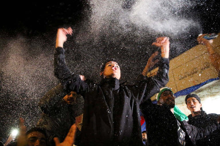 A imagem mostra um grupo de pessoas em um evento noturno， onde uma pessoa no centro está levantando os braços e parece estar se divertindo. Há uma nuvem de partículas brancas ao redor， possivelmente de confete ou espuma. As pessoas ao redor também estão animadas， e a iluminação é baixa， criando um ambiente festivo