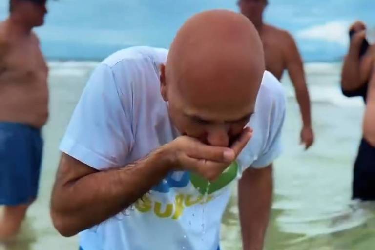 Um homem calvo， vestindo uma camiseta branca e shorts azuis， está se curvando para frente na água， aparentemente bebendo algo. Ao fundo， há outras pessoas， algumas com o torso nu， em um ambiente de praia com céu nublado.
