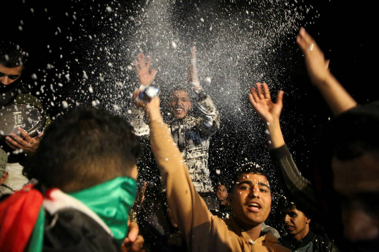 A imagem mostra um grupo de pessoas em uma celebração, com algumas delas levantando as mãos e sorrindo. Um homem no centro parece estar se divertindo, enquanto um pó branco é jogado no ar, criando um efeito festivo. Algumas pessoas estão usando máscaras ou coberturas no rosto, e há uma bandeira com as cores verde, branco e vermelho visível entre a multidão.