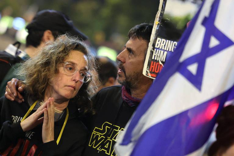 A imagem mostra um grupo de pessoas em uma manifestação. Em primeiro plano， uma mulher com cabelo cacheado e óculos， vestindo uma camiseta preta， parece preocupada e está com as mãos juntas em um gesto de oração ou reflexão. Ao lado dela， um homem com barba e cabelo escuro segura uma bandeira de Israel. Ao fundo， há outras pessoas e um cartaz que não é totalmente legível.
