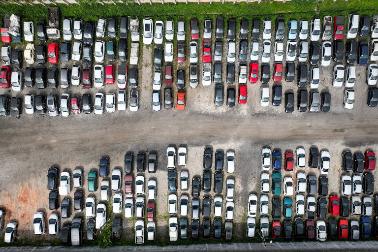 Imagem aérea de um pátio com muitos carros dispostos em filas. Os veículos são de diversas cores， incluindo branco， preto， vermelho e prata， e estão organizados em linhas bem definidas. O estacionamento é cercado por uma vegetação verde ao redor