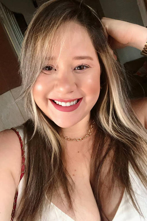 A imagem mostra uma mulher sorrindo para a câmera. Ela tem cabelo longo e liso, com mechas loiras. A mulher está usando uma blusa branca e um colar dourado. Ela segura o cabelo com uma das mãos e tem um sorriso largo, destacando seus lábios com batom. O fundo é desfocado, mas é possível ver uma parede e um sofá.