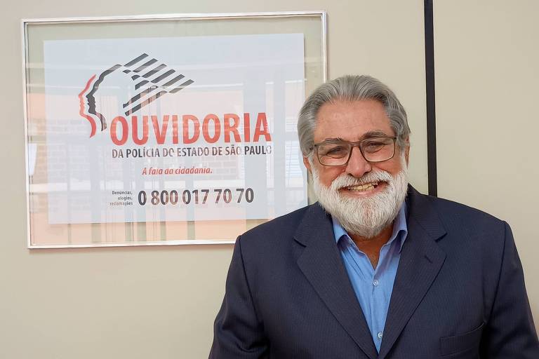 Imagem mostra um homem de cabelos grisalhos e barba， usando óculos e um terno escuro. Ele sorri em frente a um quadro que exibe informações sobre a Ouvidoria da Polícia do Estado de São Paulo. O quadro contém o texto 039;OUVIDORIA DA POLÍCIA DO ESTADO DE SÃO PAULO039;， seguido da frase 039;A fala da cidadania039; e um número de telefone 0800 017 70 70