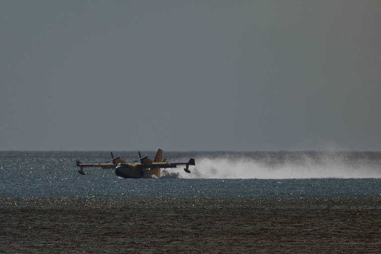 A imagem mostra um hidroavião pousando na água， levantando uma nuvem de spray. O fundo é um céu claro， e a superfície da água reflete a luz. O avião está em movimento， com as asas estendidas e a parte inferior parcialmente submersa.