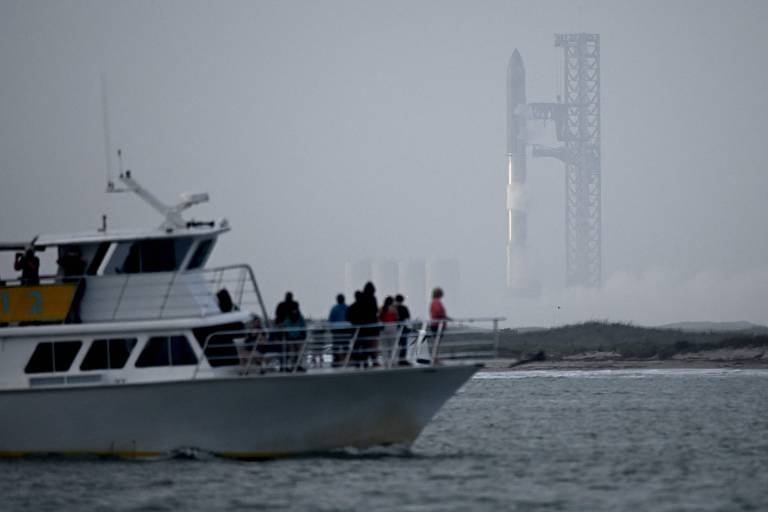 A imagem mostra um barco de passageiros navegando em águas calmas, com várias pessoas a bordo observando. Ao fundo, um foguete está posicionado em uma plataforma de lançamento, parcialmente coberto por névoa. O ambiente é nublado, criando uma atmosfera de expectativa para o lançamento.