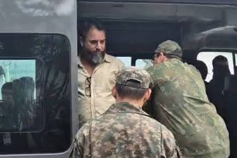 A imagem mostra um grupo de homens， alguns em uniformes militares， interagindo em torno de um veículo militar cinza. Um homem está parcialmente dentro do veículo， enquanto outro se inclina para dentro. O ambiente parece ser ao ar livre， com um céu claro ao fundo.