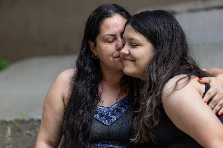 A imagem mostra duas mulheres se abraçando de forma carinhosa. Ambas têm cabelos longos e escuros. A mulher à esquerda usa uma blusa preta com detalhes azuis， enquanto a mulher à direita está vestindo uma blusa preta. Elas estão sorrindo suavemente， com expressões de afeto. O fundo é desfocado， mas parece ser uma área externa.
