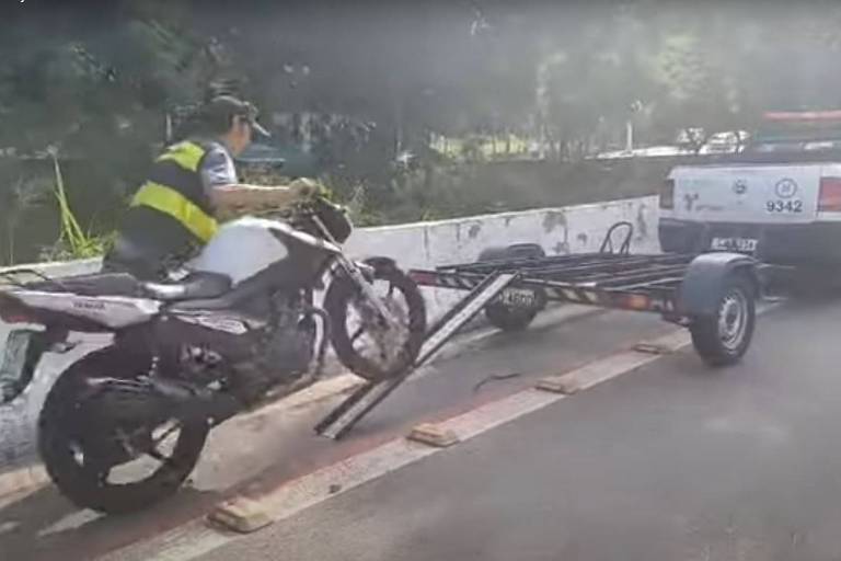 A imagem mostra um motociclista em uma motocicleta branca， que está sendo carregada por um reboque conectado a uma caminhonete. O motociclista está em pé sobre a motocicleta， enquanto a parte traseira da moto está apoiada no reboque. O cenário é uma estrada com vegetação ao fundo e uma barreira de concreto ao lado.