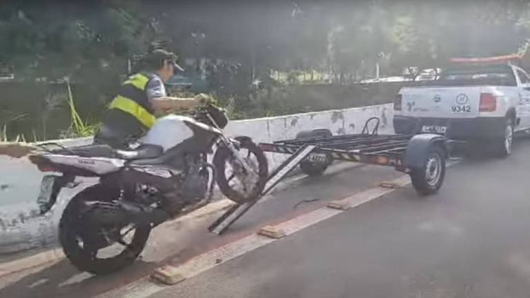 A imagem mostra um motociclista em uma motocicleta branca， que está sendo carregada por um reboque conectado a uma caminhonete. O motociclista está em pé sobre a motocicleta， enquanto a parte traseira da moto está apoiada no reboque. O cenário é uma estrada com vegetação ao fundo e uma barreira de concreto ao lado.