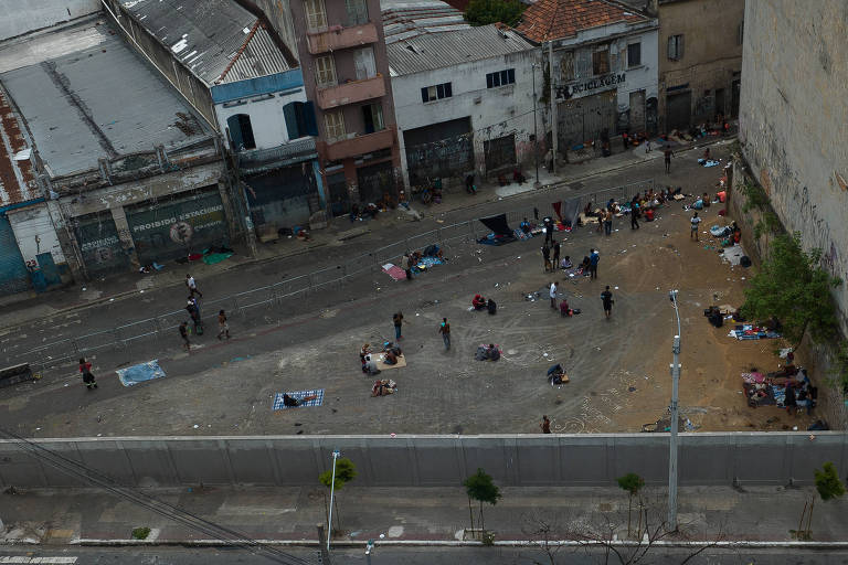 A imagem mostra uma vista aérea de uma área urbana， onde várias pessoas estão reunidas em um espaço aberto. Ao fundo， há prédios e estruturas com telhados de metal. O chão é de cimento e algumas pessoas estão sentadas ou em pé， enquanto outras parecem estar interagindo. Há também algumas árvores e um muro ao redor da área.
