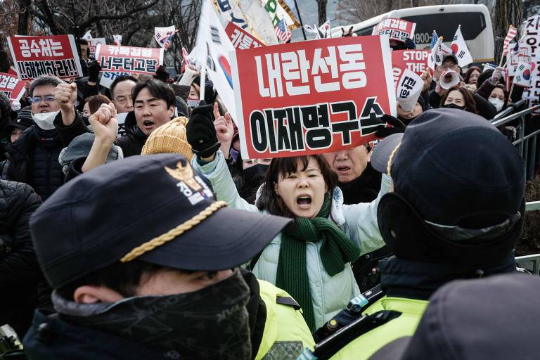 A imagem mostra um grupo de pessoas participando de um protesto. Algumas pessoas estão segurando cartazes， incluindo um que diz 039;내란선동 이재명구속039;， enquanto outras levantam os punhos e gritam. Há uma presença policial visível， com oficiais em uniformes e capacetes， que estão em frente aos manifestantes. O ambiente parece tenso， com muitos participantes usando máscaras.