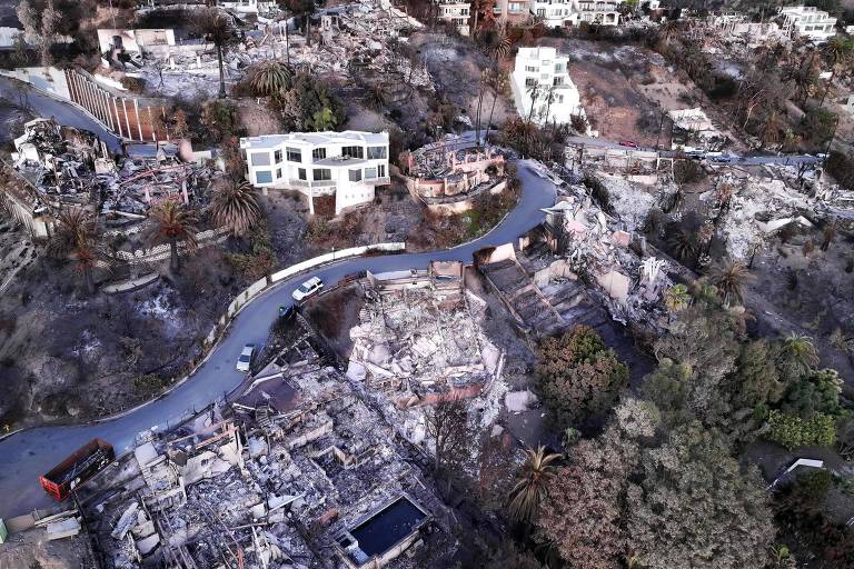 A imagem mostra uma vista aérea de uma área residencial devastada， com várias casas destruídas e terrenos queimados. Uma estrada sinuosa passa pela área， e algumas casas ainda permanecem de pé， contrastando com os escombros ao redor. A vegetação está danificada， e o cenário transmite uma sensação de destruição e abandono.