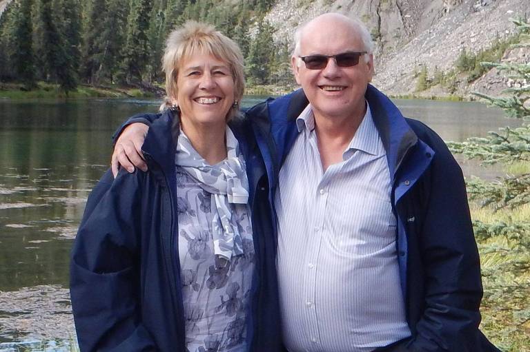 Liz Barlow e Ivan Greenaway estão abraçados em frente a um lago， com roupas de frio. A mulher usa uma blusa clara com um padrão e um casaco escuro， enquanto o homem está vestido com uma camisa clara e um casaco escuro. Ambos parecem felizes e estão próximos um do outro.
