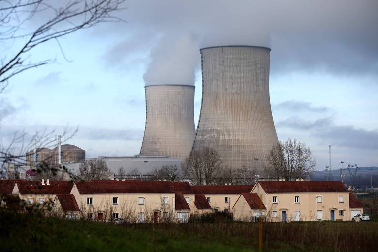 A imagem mostra duas torres de resfriamento de uma usina nuclear， com fumaça saindo de suas partes superiores. Ao fundo， há um céu nublado e， na parte inferior da imagem， estão visíveis algumas casas de um bairro próximo à usina.