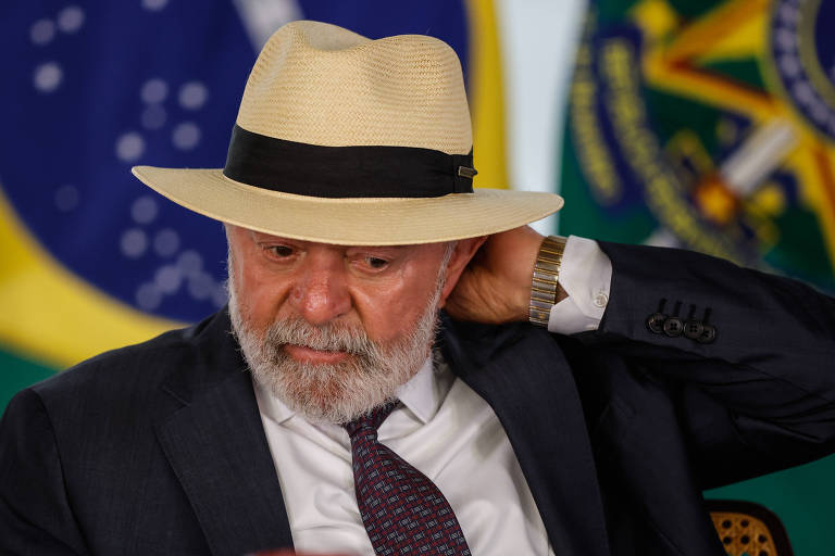 Imagem mostra Lula, um homem de barba e cabelo grisalho, vestindo um terno escuro e uma gravata, e que está sentado em um evento. Ele usa um chapéu claro com uma faixa preta e parece estar ajustando o chapéu com a mão. Ao fundo, há bandeiras do Brasil.