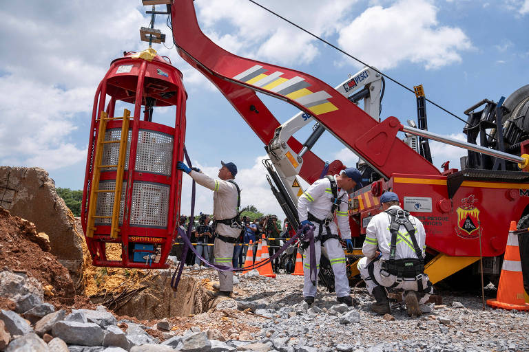 A imagem mostra uma equipe de resgate trabalhando em uma operação. Um guindaste vermelho está levantando uma cabine de resgate， enquanto vários membros da equipe， vestidos com roupas brancas e capacetes， estão ao redor， observando e ajudando. O cenário é ao ar livre， com céu parcialmente nublado e cones de sinalização laranja ao fundo