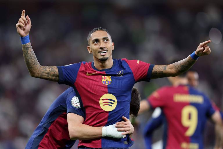 Com os braços abertos， sorrindo e abraçado na altura da cintura por um colega de Barcelona， Raphinha festeja gol na decisão da Supercopa da Espanha， em Jedá， na Arábia Saudita， contra o Real Madrid