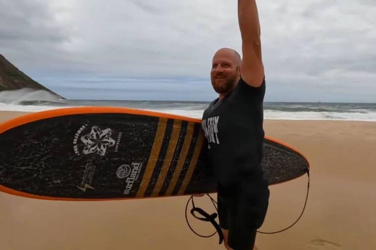 Um homem sorridente está em uma praia segurando uma prancha de surf com uma mão levantada em sinal de vitória. Ele usa uma camiseta preta e shorts. Ao fundo， há ondas do mar e uma colina. O céu está nublado.