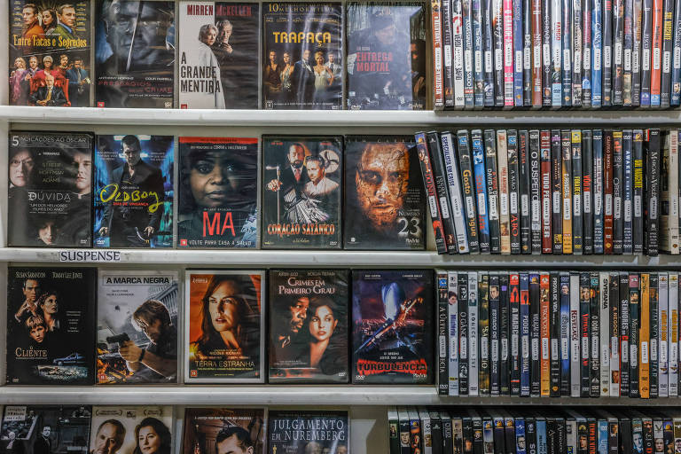 A imagem mostra uma prateleira cheia de DVDs organizados em várias fileiras. Na parte inferior, há capas de filmes com imagens de personagens e títulos visíveis. Na parte superior, há uma coleção de DVDs em caixas mais simples, organizados verticalmente. A prateleira é de cor clara e os DVDs estão dispostos de forma ordenada.