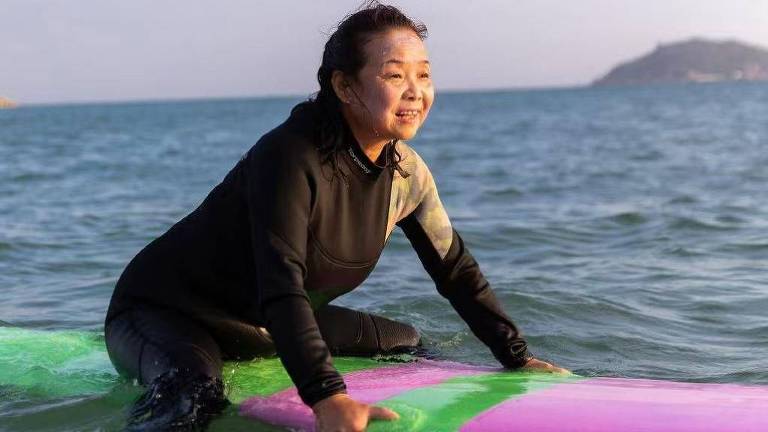 Uma mulher está em cima de uma prancha de surf colorida， com um traje de neoprene preto. Ela sorri enquanto se equilibra na prancha， que é verde e rosa， em águas calmas. O fundo mostra o mar e uma costa ao longe， sob a luz suave do sol poente.