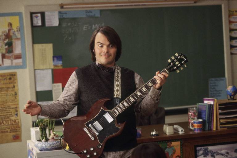 Um homem com cabelo escuro e liso， usando um colete cinza e uma camisa com estampa de bolinhas， está em pé em uma sala de aula. Ele segura uma guitarra elétrica marrom e faz uma expressão animada. Ao fundo， há uma lousa verde e vários cartazes e materiais escolares nas paredes. Em uma mesa próxima， há livros， um globo terrestre e um recipiente com doces.