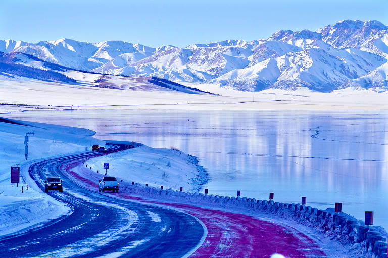 A imagem mostra uma estrada sinuosa coberta de neve， com um lago congelado à direita e montanhas ao fundo. O cenário é predominantemente azul， refletindo a luz do céu e a neve. Não há pessoas visíveis na imagem.