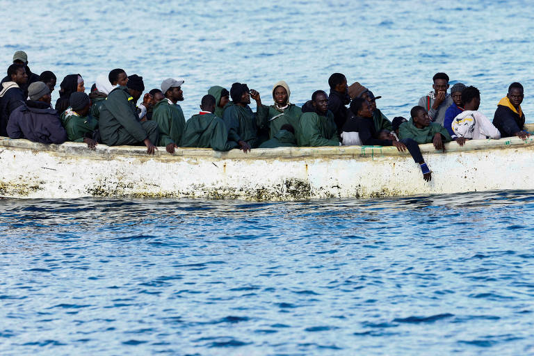 Um grupo de pessoas está sentado em uma embarcação pequena， flutuando em águas azuis. A maioria das pessoas está vestida com capas verdes， e algumas estão usando bonés ou chapéus. O ambiente parece ser ao ar livre， com um céu claro ao fundo.