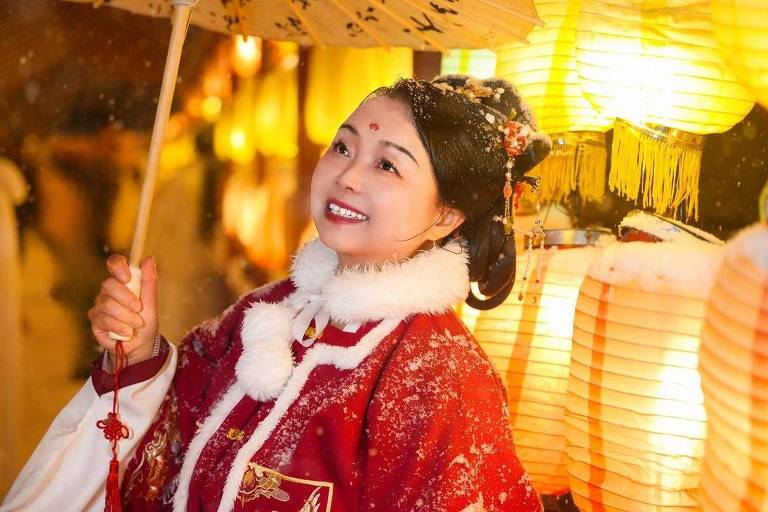 Su Min aparece vestindo um traje tradicional chinês. Ela está de pé， usando um vestido vermelho com gola de pele na frente de várias lâmpadas de papel amarelas acesas.