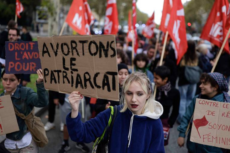 A imagem mostra uma manifestação com várias pessoas segurando bandeiras vermelhas e cartazes. Em primeiro plano， uma jovem com cabelo claro e roupas escuras segura um cartaz que diz 039;AVORTONS LE PATRIACT039;. Ao fundo， outras pessoas também estão segurando cartazes e bandeiras， criando um ambiente de protesto