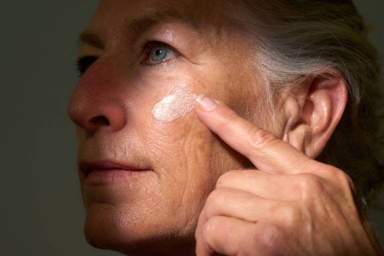 A imagem mostra uma mulher de pele madura aplicando creme no rosto. Ela utiliza o dedo indicador para espalhar o produto， evidenciando rugas e linhas de expressão características da idade. A iluminação destaca parte de seu rosto， enquanto o fundo é escuro， dando foco à ação de cuidado com a pele. A composição parece transmitir a ideia de autocuidado e cuidados com o envelhecimento da pele.