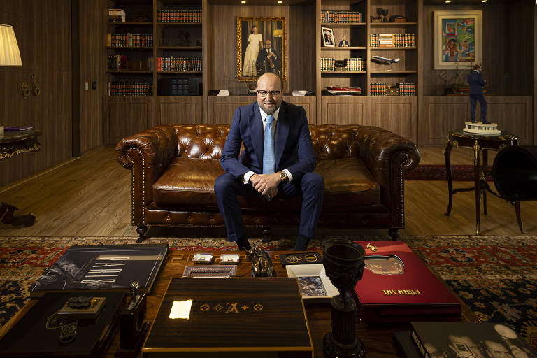 Um homem sentado em um sofá de couro marrom em um escritório bem decorado. Ele usa um terno escuro e óculos. À sua frente， uma mesa com vários objetos， incluindo pastas e livros. Ao fundo， uma estante com livros e obras de arte nas paredes.