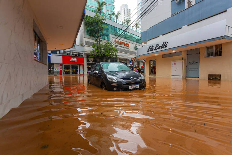 A imagem mostra uma rua inundada em uma área urbana， com água cobrindo o chão. Um carro preto está parado em meio à água， enquanto prédios e lojas são visíveis ao fundo. A água parece ter uma coloração alaranjada， possivelmente devido a sedimentos ou poluição. O clima parece nublado， sugerindo que a chuva pode ter causado a inundação.