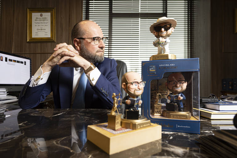 Um homem de óculos e terno azul está sentado em uma mesa de escritório, com as mãos entrelaçadas. Ao seu lado, há três estatuetas em uma mesa de mármore, incluindo uma em uma caixa. O ambiente é bem iluminado, com janelas ao fundo e documentos organizados na mesa.