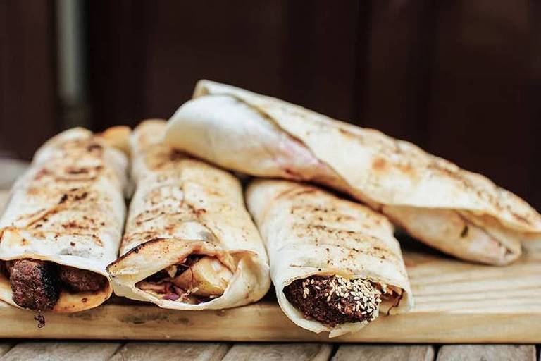 A imagem mostra quatro wraps grelhados dispostos em uma tábua de madeira. Os wraps são enrolados e apresentam diferentes recheios visíveis nas extremidades, incluindo carne e vegetais. A superfície dos wraps tem marcas de grelha, indicando que foram cozidos em uma grelha.