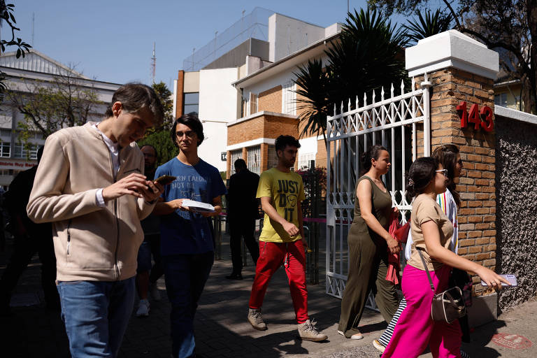 A imagem mostra um grupo de pessoas caminhando em frente a um edifício. Algumas pessoas estão paradas， enquanto outras estão em movimento. Um homem à esquerda está segurando um prato de comida. O ambiente é urbano， com um céu claro e edifícios ao fundo. As pessoas vestem roupas de diferentes cores e estilos.